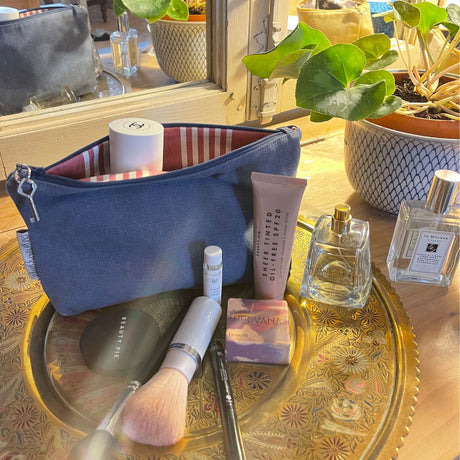 A medium-sized blue zipped pouch used as a makeup bag, displayed on a table with various toiletry items like perfume bottles, makeup brushes, and lotion tubes arranged around it.