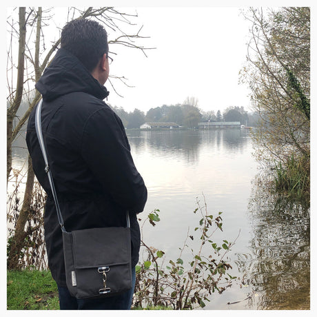 A person standing by a body of water wearing a black coat and carrying a silver-grey cross body bag with a black clip closure.