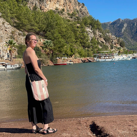 A person standing by a body of water wearing a striped handbag in a crossbody style with a mountainous backdrop.
