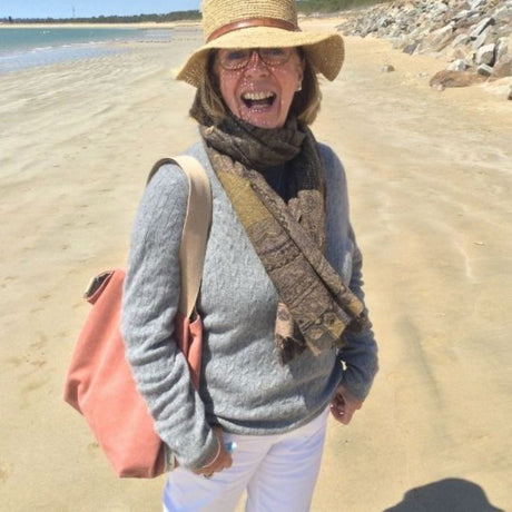 An older woman standing on a beach wearing a wide-brimmed hat and carrying a tote bag with thick shoulder straps.
