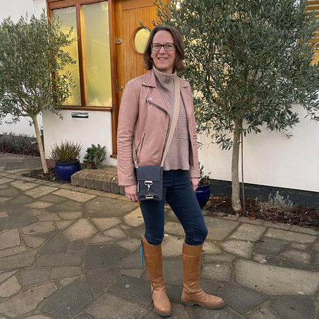 A person wearing a pink jacket and blue jeans, standing on a pathway with trees and a house in the background, carrying a compact blue waterproof crossbody bag.