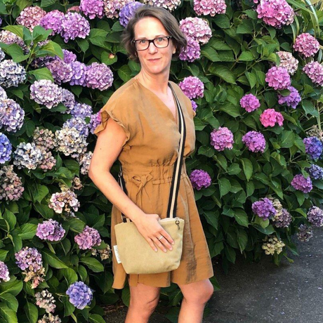 A person wearing a beige crossbody bag with a shoulder strap, standing in front of a wall of flowers.